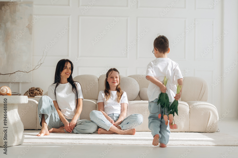 Fototapeta premium Woman and two children sitting on a light-colored rug in a modern living room, child playfully holding green leaves behind back, soft natural light illuminating the scene
