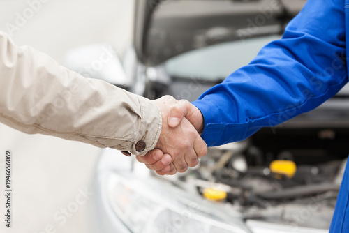 Close-up of a customer and a professional auto mechanic shaking hands in front of a broken car with an open hood.
