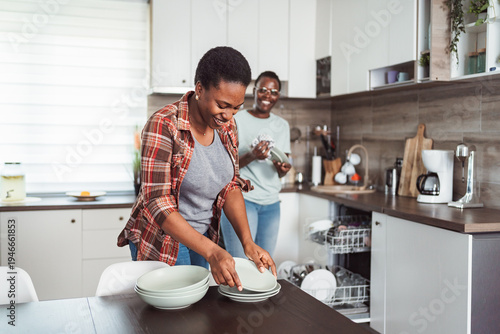 Картината върху платно Two cheerful women putting away clean dishes after unloading dishwasher in moder