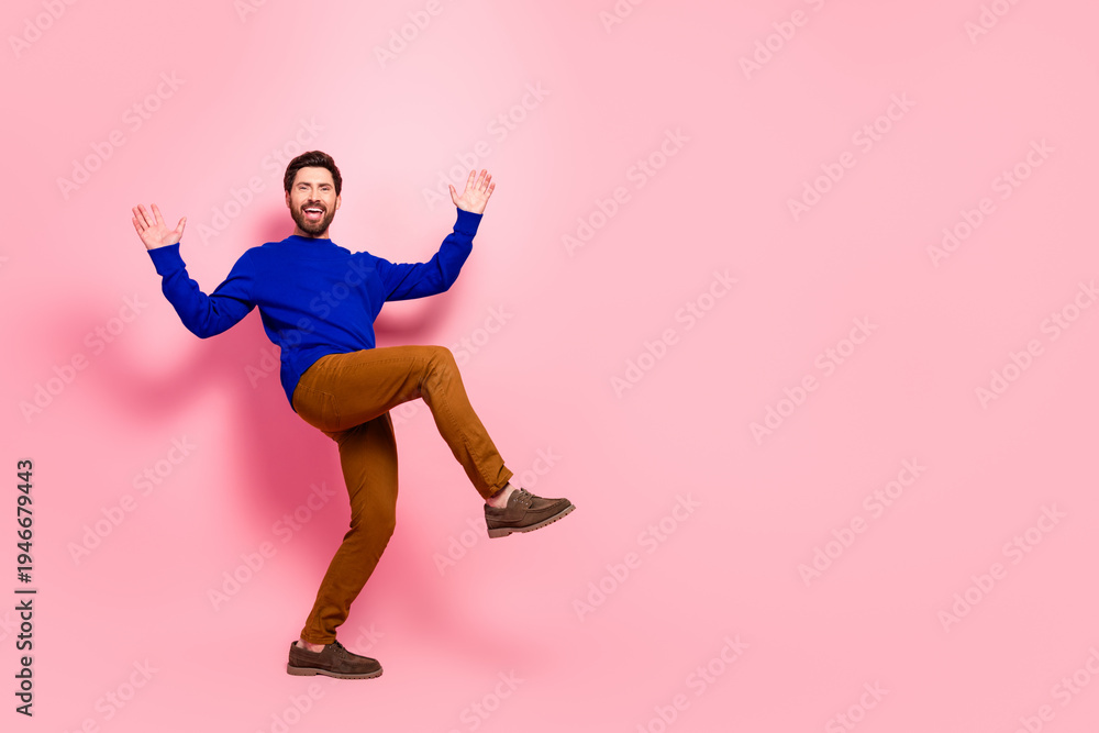Obraz premium Happy young man in blue sweater and brown pants leaps with arms raised against pink background expressing energy joy and a casual lifestyle for fashion advertising