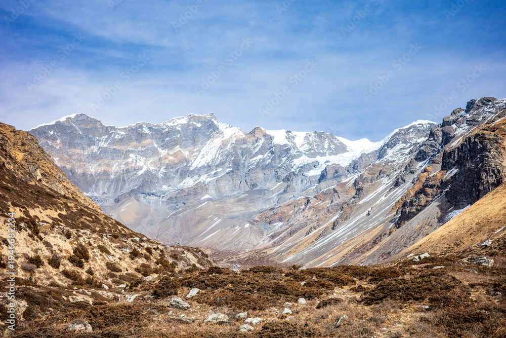 Obraz premium Wide panoramic view of the Himalayan mountain range featuring towering snow-capped peaks rising above a rugged alpine valley. The dramatic landscape showcases steep rocky slopes