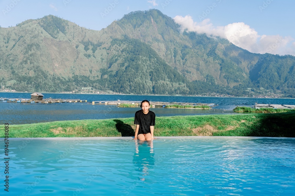 Fototapeta premium Woman in black swimsuit standing in pool with mountain and lake background