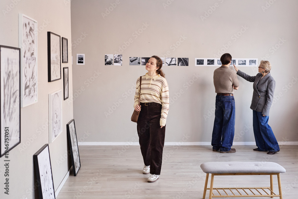 Fototapeta premium Young adult Caucasian woman walking through art gallery, middle aged Caucasian man and senior woman examining black and white photographs displayed on wall