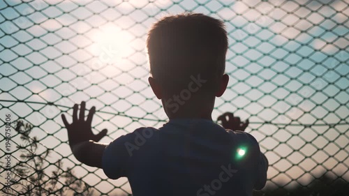 Boy reaching hands through chainlink fence toward sunlight outdoors. Child raises arms against fence barrier. Silhouette boy reaches through chainlink. Child lifts hands at wire fence with sunlight.