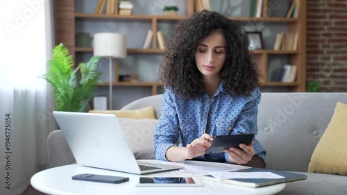 Young busy woman dealing with household finances sitting on sofa in living room at home office. Beautiful female doing paperwork, calculates on a calculator, writes down data in document, uses laptop
