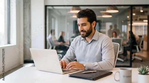 A man working on his laptop in a modern office environment with colleagues in the background