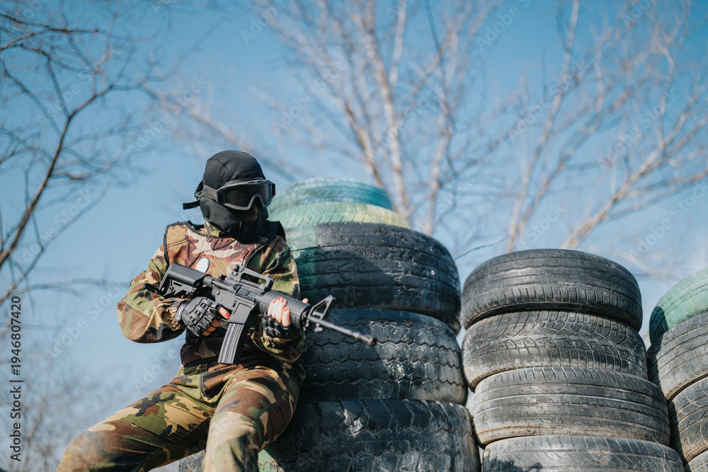 Fototapeta premium Outdoor paintball scene featuring a camo-clad player with protective mask and rifle at tire cover.