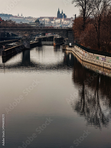 Historic city bridge over calm river with reflection and old european architecture skyline in background, urban landscape with river Vltava. Prague