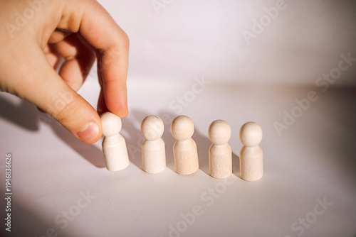Hand placing wooden figurine among four others on a light surface, showcasing a simple arrangement of minimalistic toys or game pieces in a studio setting