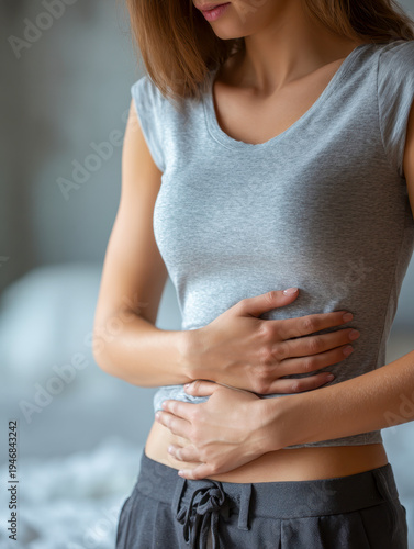 Woman in casual wear holding her stomach as she experiences abdominal discomfort or pain indoors with a blurred background suggesting a home environment