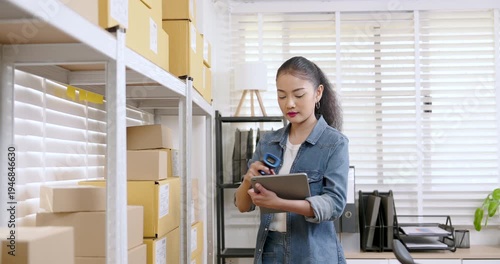 Asian woman small business owner slowly reviews inventory list on tablet while standing between warehouse shelves filled with online shopping parcel boxes ready for shipment
