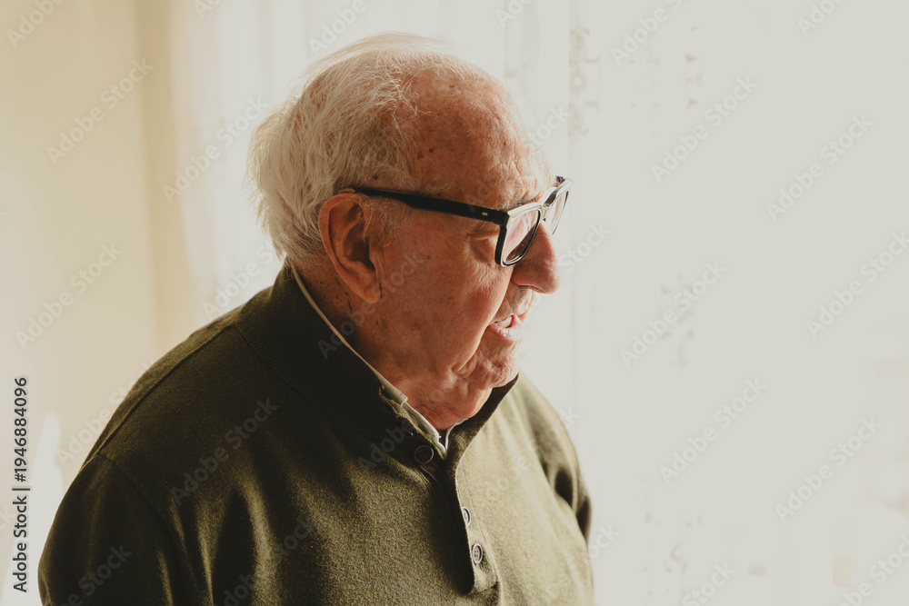 Fototapeta premium Elderly man with glasses looking through window at home