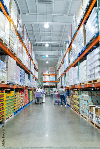 A wide interior photograph of a large warehouse-style wholesale shopping center featuring long aisles lined with tall pallet racks filled with bulk merchandise. The image captures the spacious retail 
