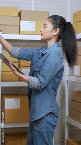 Asian woman small business owner holding tablet while pointing finger upward, checking inventory location, managing online shopping order workflow, coordinating e-commerce stock inside home warehouse