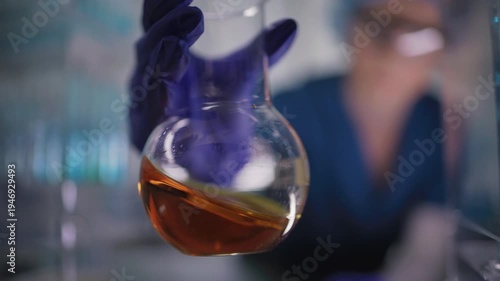 Female researcher preparing a new type of enriched bio fuel. Holding a glass flask with orange liquid close to the camera. Selective focus