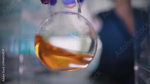 Female researcher preparing a new type of enriched fuel. Holding a glass flask with orange liquid close to the camera and swirling the liquid. Selective focus
