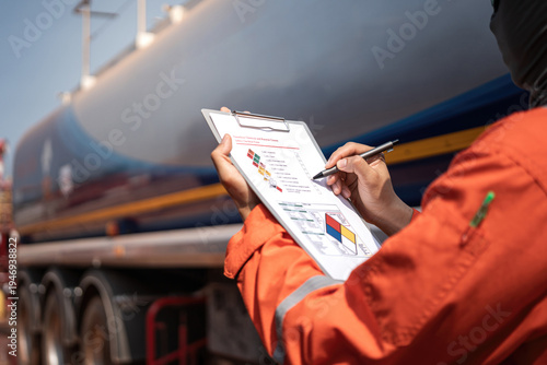 An engineer is checking on chemical hazardous material checklist to verify the safety condition with chemical road tanker as background. Industrial waorking scene, close-up and selective focus.
