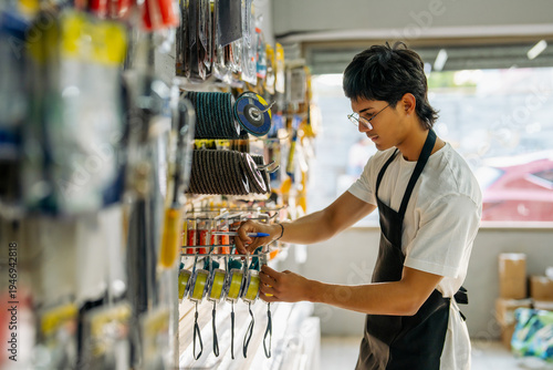 Photos Young man wearing apron and glasses organizing products on a shelf in a hardware