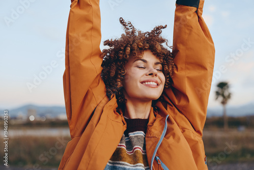 Happy woman with curly hair wearing rainbow sweater and orange jacket smiles with eyes closed stretching arms outdoors in nature, enjoying lifestyle moment with positive mood and smartphone.