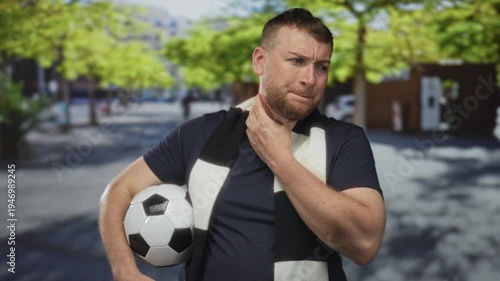 Young heavyset man holding soccer ball under arm and clutching neck with hand on busy street in front of building, wearing black and white scarf and navy t shirt; discomfort.