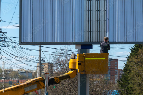 Worker on Hydraulic Lift Maintaining Large Billboard Sign by Road Under Power Lines in Urban Area