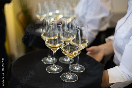 Close-up of a server holding a tray with several glasses of white wine in a restaurant or event setting. Soft background blur with additional glasses suggests a formal dining or reception atmosphere