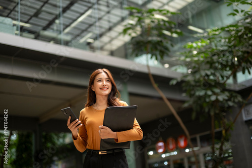 Young businesswoman holding phone and folder walking in office lobby