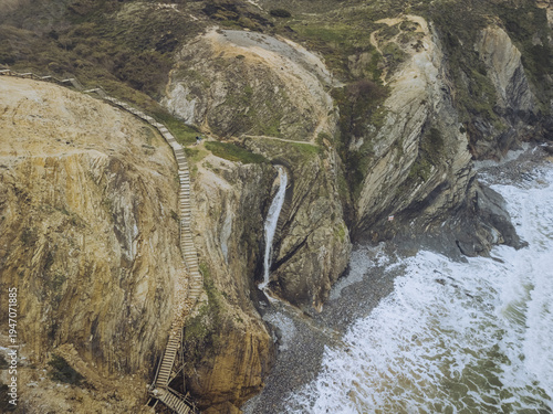 Aerial view of a cascading waterfall plunging onto a rugged beach where the cliffs meet the restless ocean, Zambujeira do Mar, Beja District, Portugal.