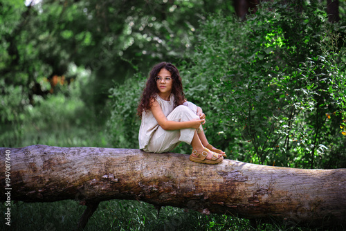 Little girl with curly hair sitting on a fallen tree trunk in a summer forest