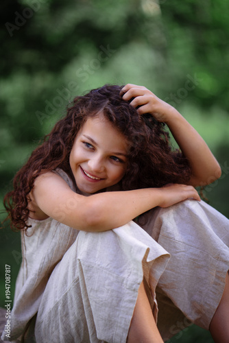 Adorable young girl with curly hair standing by a large tree in a green summer forest