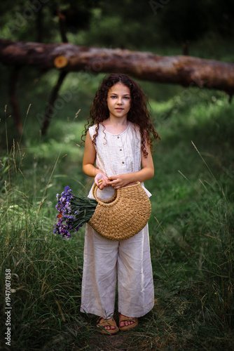 Little girl with long curly hair holding a woven straw bag with purple flowers in a forest path