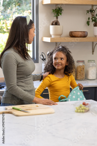 Diverse mother placing hand on child's shoulder at counter by window, child holding blue backpack