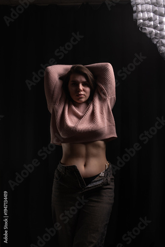 Female model with long dark hair poses in a pink ribbed sweater and high-waisted jeans against a dark backdrop, illuminated by soft overhead lighting