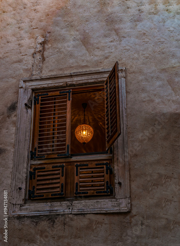 Croatia, Europe: crystal chandelier with the light on glimpsed from a wooden shuttered window in the alleys of the old town of Split. Stone wall, ancient building 
