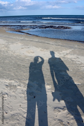 Shadows of two people on sandy beach. Long shadows of two people on a sandy beach near the sea on a sunny day. Coastal landscape with waves and blue sky creating a relaxed vacation and summer atmosphe