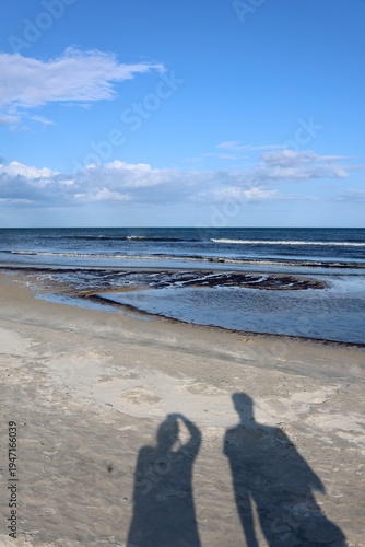 Shadows of two people on sandy beach by the sea. Long shadows of two people on a sandy beach near the sea on a sunny day. Coastal landscape with waves and blue sky creating a relaxed vacation