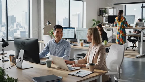Two colleagues collaborating at their desks in a modern office environment, discussing work on computer screens and laptops