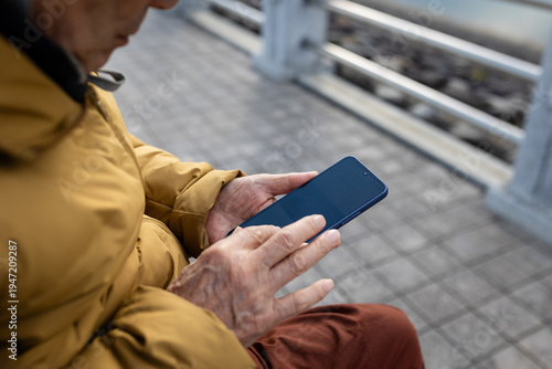 An older man sitting on a bench by the lakeside, looking at his mobile phone