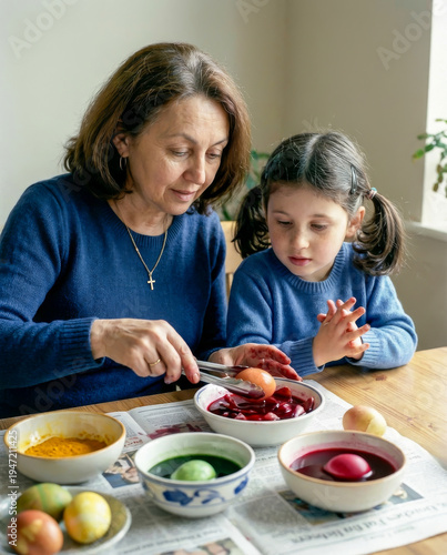 Grandmother and granddaughter color Easter eggs with bright dye at wooden table indoors.