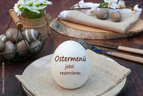 Osterei mit der Aufschrift 'Ostermenü jetzt reservieren' auf einem Teller mit Stoffserviette vor einem Korb mit bemalten Ostereiern und einem Holzbrett mit Besteck