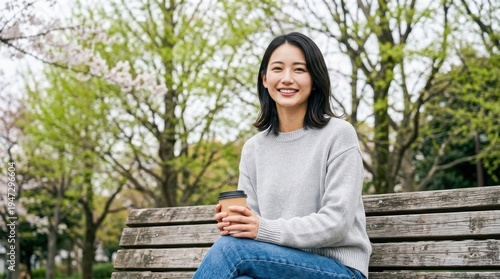公園のベンチに座りコーヒーを片手に微笑む若い女性