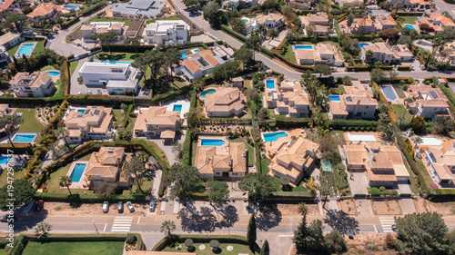 Aerial view shows many houses with swimming pools surrounded by trees and roads in a residential area on a sunny day. People enjoy outdoor activities