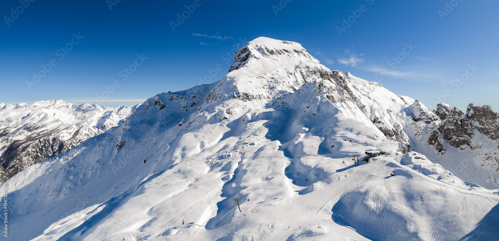 Fototapeta premium Aerial view shows San Domenico ski resort in the Italian Alps. Groomed pistes line a snow peak, ski lifts and a small station sit on a ridge, skiers descend broad slopes.
