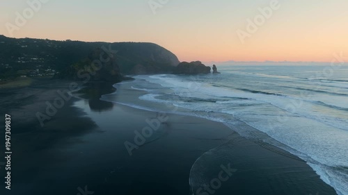 Aerial view of Piha Beach, New Zealand, at dusk. The waves are crashing on the shore, and the sky is a beautiful orange color. Lion Rock is a popular tourist destination.