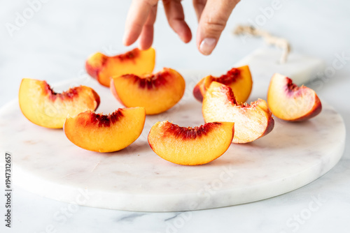 Fotografie Sliced segments of free stone peaches on a marble slab with a hand reaching in