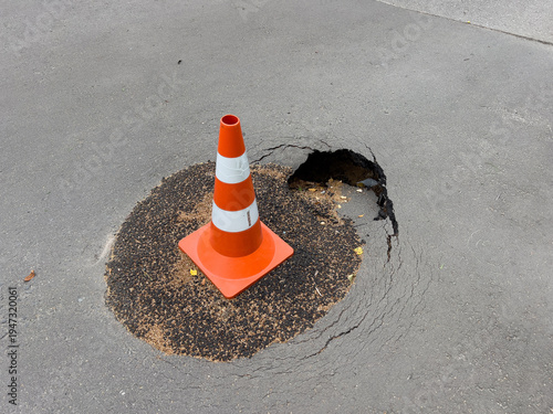 Orange traffic cone standing next to a severe pothole in cracked asphalt, indicating road hazard and public safety risk