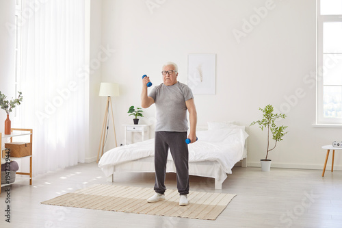 Tableau sur toile Focused gray haired senior man standing in bedroom and lifting dumbbells
