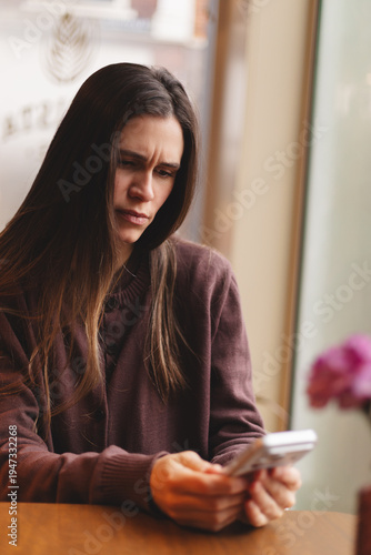 Concentrated and worried young woman looking at smartphone screen with frowning face, wearing burgundy cardigan in a cafe, confused girl reading bad news or having problem with mobile phone app