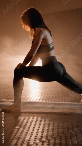 Focused female athlete stretches in a modern yoga studio, illuminated by a glowing light source behind her. The serene atmosphere and mist create a tranquil, inspiring fitness scene