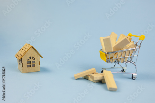 small home shopping, miniature dwelling beside shopping trolley with scattered wooden pieces, scale model house positioned near shopping cart filled with wooden fragments indicating